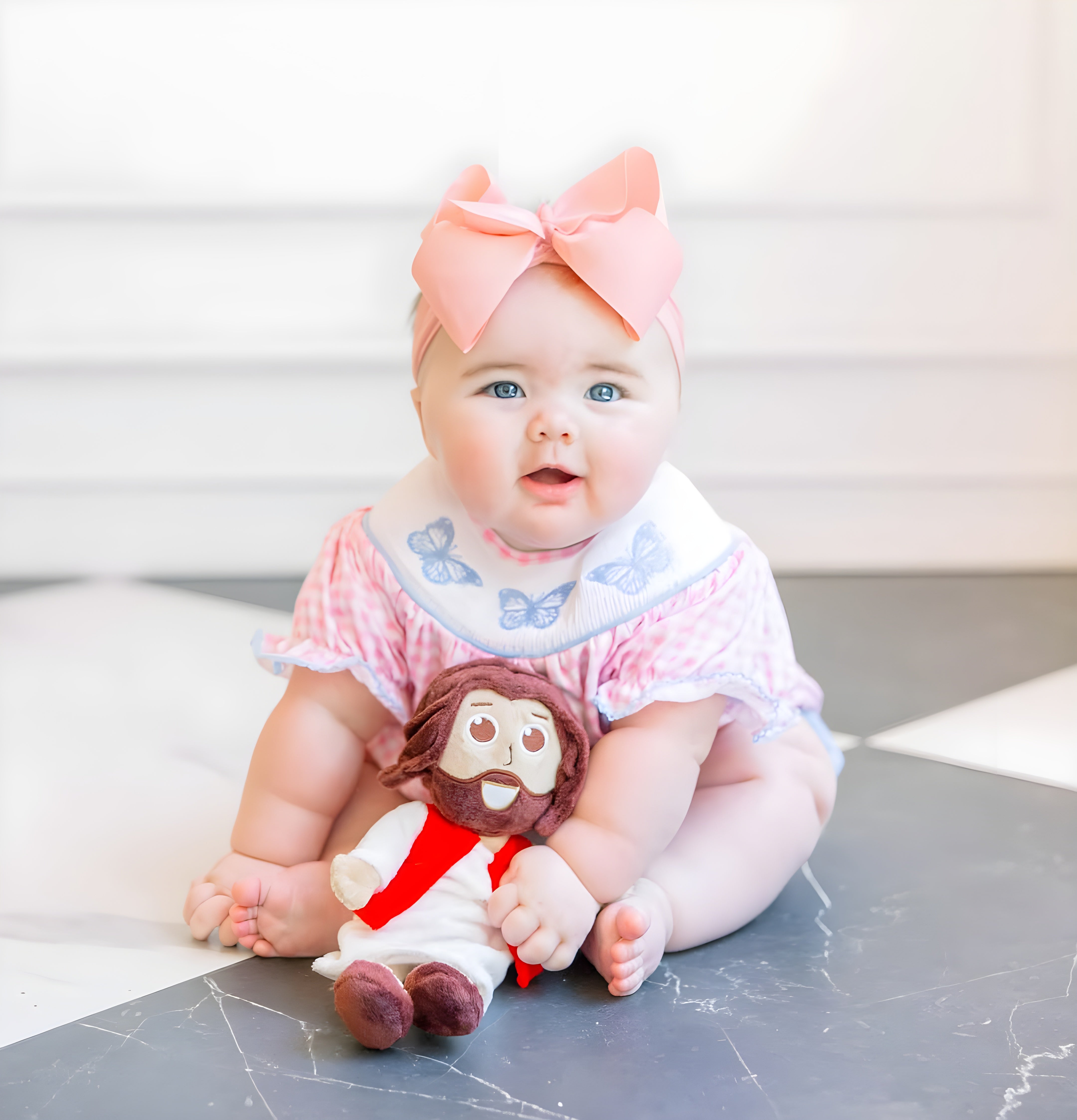A baby wearing a pink and white ruffled outfit with a large pink bow headband and a white bib with blue butterflies, sitting on a dark marble floor and holding a lil' jesus doll from jesusdoll.com.  courtesy everleighelaine.com